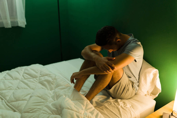 Depressed hispanic man in pajamas hugging his knees and feeling lonely in bed at midnight - Photo, Image