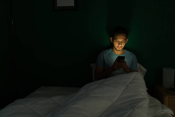 Ich kann nicht schlafen. Attraktiver Mann sitzt mitten in der Nacht in seinem Bett, während er SMS schreibt und sein Smartphone benutzt - Foto, Bild