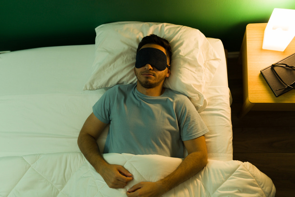 Δεν μπορώ να κοιμηθώ με τα φώτα αναμμένα. Υψηλή γωνία ενός όμορφου άντρα με μάσκα ύπνου ξαπλωμένος στο άνετο κρεβάτι του - Φωτογραφία, εικόνα