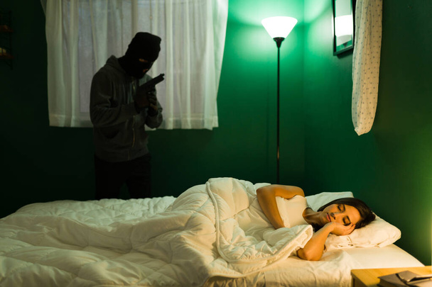Thief with a mask and a gun committing a robbery at a home while a young woman sleeping in the bedroom - Foto, immagini