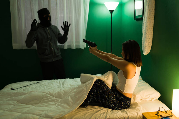 Get out of my house. Scared woman in bed pointing a gun to a male thief breaking in during the night - Photo, Image