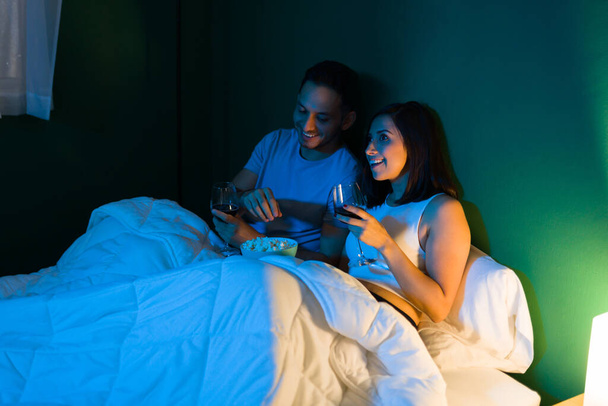 Piękna para ogląda razem film w łóżku przed pójściem spać jedząc popcorn i pijąc wino  - Zdjęcie, obraz