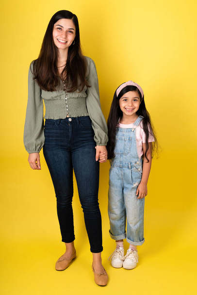 Jolie jeune mère et mignonne petite fille tenant la main tout en établissant un contact visuel sur un fond de studio jaune - Photo, image