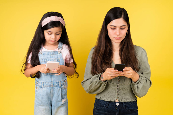 Привлекательная молодая женщина и маленькая девочка используют смартфоны, отправляя смс на желтый фон - Фото, изображение