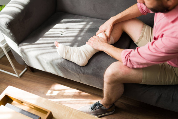 Vista superior de un joven en el sofá recuperándose de una lesión en la pierna y el pie  - Foto, imagen