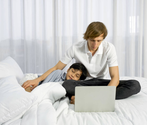 父は息子と一緒に家で働いています。父親がラップトップコンピュータを使って寝室で働いている間、父親の膝の上で昼寝をしている少年。ワークライフバランス.  - 写真・画像