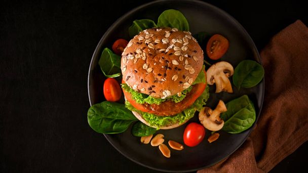 Vista superior, vista superior de un plato de deliciosa hamburguesa vegana con pan de grano entero, carne vegana y verduras orgánicas frescas sobre fondo negro. - Foto, Imagen