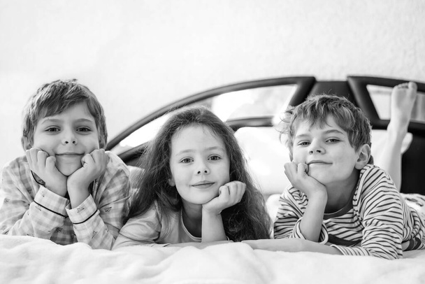 Tres niños felices en pijama celebrando la fiesta de pijama. Niños y niñas preescolares y escolares en ropa de dormir divirtiéndose juntos. Niños jugando juntos en la cama. Haciendo pelea de almohadas, en el interior de casa. - Foto, Imagen