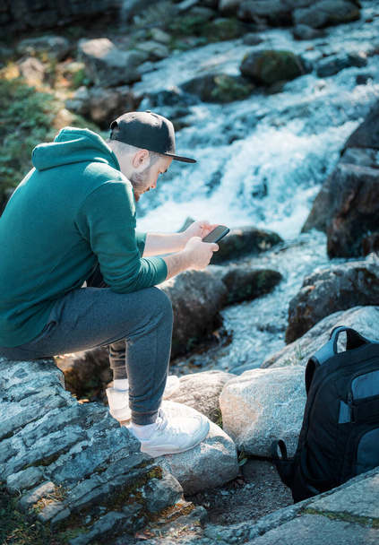 Yürüyüş sırasında dinlenen bir adam kayaya oturmuş telefonunu karıştırıyor. - Fotoğraf, Görsel