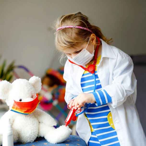 Une petite fille fait une injection à un ours en peluche. Enfant mignon avec masque médical jouant le médecin, tenant la seringue avec le vaccin. Coronavirus covid vaccination concept. Jeu de rôle pour enfants à la maison. - Photo, image