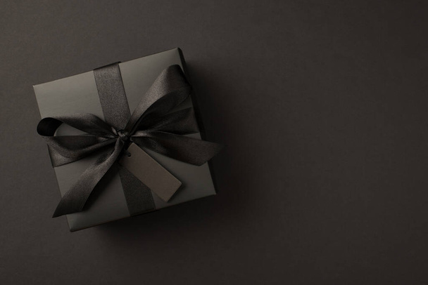 Top view photo of giftbox in black packaging with black satin ribbon bow and tag on isolated black background with copyspace - Foto, Imagen