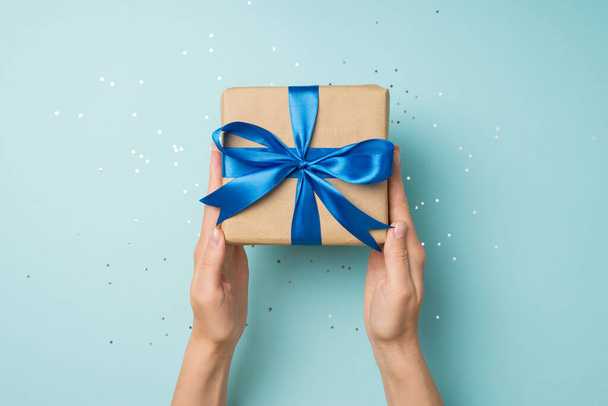 First person top view photo of woman's hands holding craft paper giftbox with blue ribbon bow over shiny confetti on isolated pastel blue background - Φωτογραφία, εικόνα