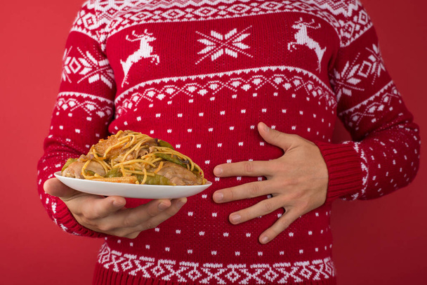 Обрізаний крупним планом фотографія людини в червоно-білому різдвяному светрі, що тримає велику тарілку їжі і торкається його болючого шлунка на ізольованому червоному тлі
 - Фото, зображення