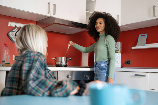 Two young multiracial students girls cooking in the youth hostel kitchen talking an socializing together - 写真・画像
