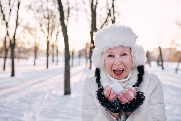 vanhempi nainen valkoisessa hatussa ja turkissa pitämässä lunta käsissä lumimetsässä. Talvi, ikä, kausi käsite  - Valokuva, kuva