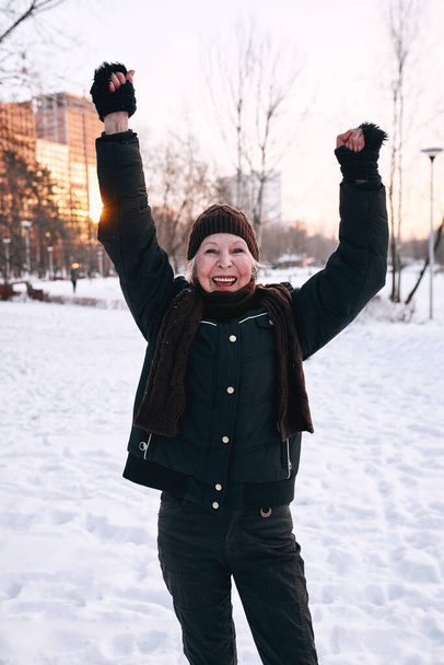 ηλικιωμένη γυναίκα με καπέλο και σπορ μπουφάν που κάνει τζόγκινγκ στο χειμερινό πάρκο. Χειμώνας, ηλικία, αθλητισμός, δραστηριότητα, έννοια εποχής - Φωτογραφία, εικόνα