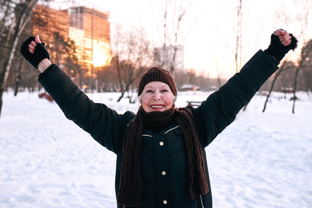 старшая женщина в шляпе и спортивной куртке бегает трусцой в зимнем парке. Зима, возраст, спорт, активность, концепция сезона - Фото, изображение