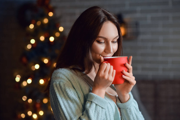Όμορφη νεαρή γυναίκα με φλιτζάνι ζεστό κακάο ποτό στο σπίτι την παραμονή των Χριστουγέννων - Φωτογραφία, εικόνα