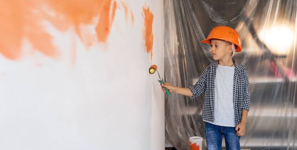 Kinder malen drinnen. Ein kleiner Junge mit orangefarbenem Helm repariert ein Haus und bemalt die Wand mit einer Walze. Das Kind hilft beim Dekorieren der Wände. Renovierung - Foto, Bild
