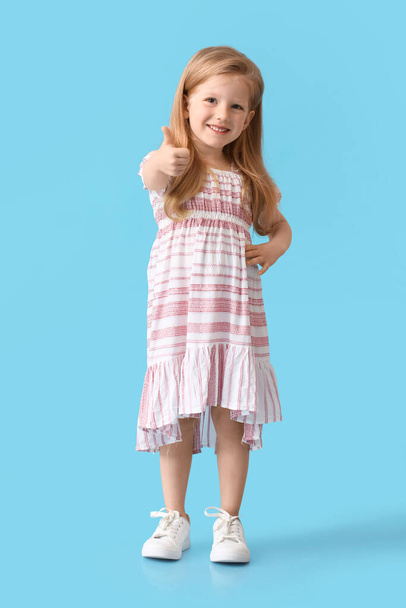 Klein meisje in schattige jurk tonen duim-up op blauwe achtergrond - Foto, afbeelding