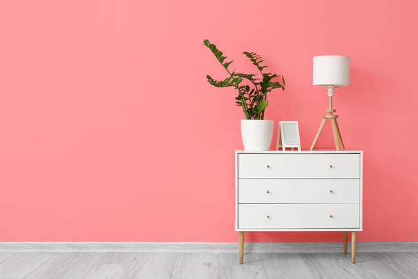Lámpara blanca con marco y planta de interior en la cómoda cerca de la pared rosa - Foto, Imagen