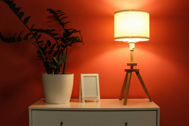 Lámpara brillante con marco y planta de interior en estante en habitación oscura - Foto, Imagen