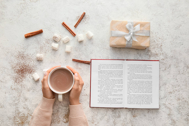 Bir fincan kakao, marşmelov, tarçın, kitap ve ışık arkaplan üzerine hediye almış kadın elleri. - Fotoğraf, Görsel