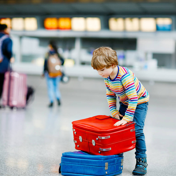 国際空港のターミナルで大きなスーツケースの荷物を持つ幸せな小さな男の子。就学前の興奮した子供は飛行機で休暇に行くのを待ちます。家族旅行の生活. - 写真・画像
