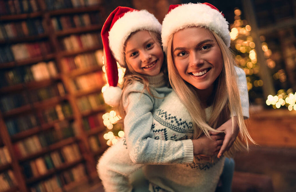 Gyönyörű anya és lánya, vannak karácsonyi kalapjai, karácsonyfás fényei. Portré ünnep. Téli háttér. Családi gyerekportré. Újévi dekoráció. - Fotó, kép
