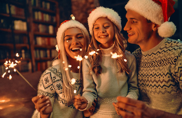 Wesołych Świąt i Szczęśliwego Nowego Roku! Szczęśliwa rodzina świętująca zimowe wakacje w domu. Rodzice i ich córka czekają na Boże Narodzenie w kapeluszach Mikołaja z ogniem w rękach. - Zdjęcie, obraz