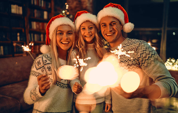 Wesołych Świąt i Szczęśliwego Nowego Roku! Szczęśliwa rodzina świętująca zimowe wakacje w domu. Rodzice i ich córka czekają na Boże Narodzenie w kapeluszach Mikołaja z ogniem w rękach. - Zdjęcie, obraz