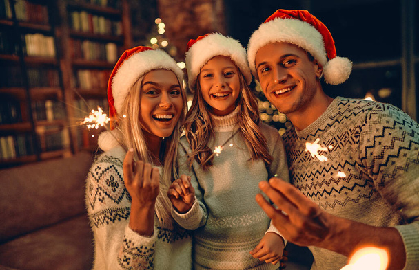 Joyeux Noël et bonne année ! Bonne famille célébrant les vacances d'hiver à la maison. Les parents et leur fille attendent Noël dans des chapeaux de Père Noël avec des étincelles dans les mains. - Photo, image