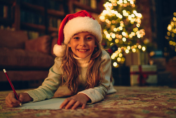 Feliz Natal e Boas Festas! Menina bonito escreve a carta para Papai Noel perto da árvore de Natal dentro de casa
. - Foto, Imagem