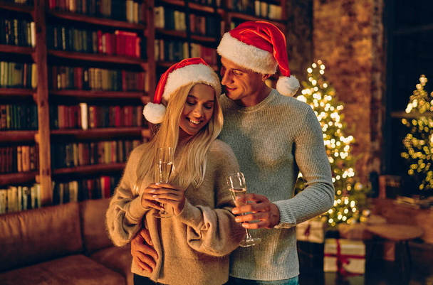 Beautiful young couple in Santa hats is holding glasses of champagne and smiling while celebrating New Year at home. Against the background of the Christmas tree. - Foto, immagini