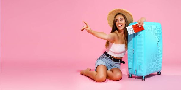 Glad girl with suitcase, tickets, money and passport are going to travel while pointing finger - Фото, изображение