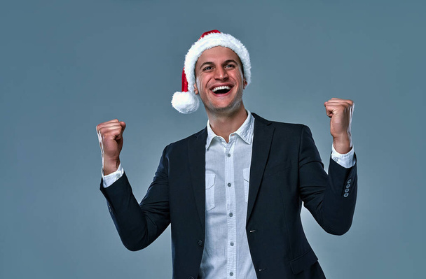 Attractive successful businessman in a jacket and shirt in a cap of Santa Claus say yes in the studio on a gray background. - Foto, Imagem