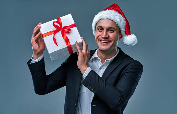 Привабливий успішний бізнесмен в піджаку і сорочці в кепці Санта Клауса з подарунковою коробкою в студії на сірому фоні. - Фото, зображення