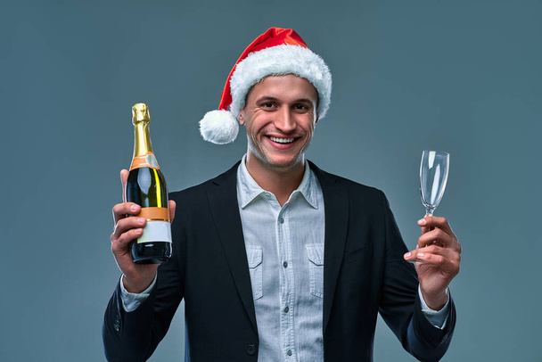 Un hombre exitoso con chaqueta y sombrero de Navidad con una botella de champán celebra el Año Nuevo. Foto del estudio sobre un fondo gris . - Foto, Imagen
