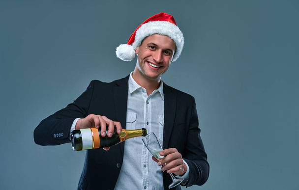 Successful man in a jacket and Christmas hat pours champagne into a glass celebrates New Year. Studio photo on a gray background . - Valokuva, kuva