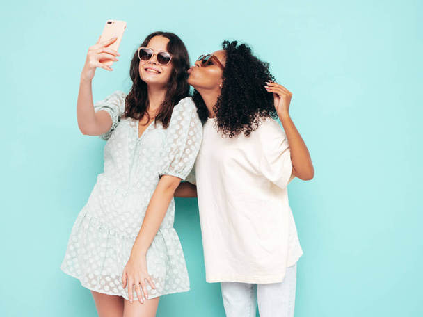 Kaksi nuorta kaunista hymyilevää kansainvälistä hipsterinaista trendikkäissä kesävaatteissa. Seksikäs huoleton naiset poseeraa lähellä sinistä seinää studiossa. Positiiviset mallit hauskaa aurinkolasit. Valokuvien ottaminen selfiestä - Valokuva, kuva