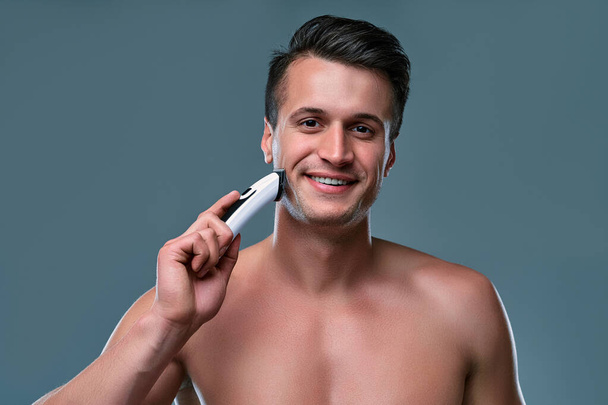 Jóképű fiatalember elszigetelve. Póló nélküli izmos férfi portréja szürke háttéren áll, borotválkozás közben vágóval a kezében. Férfi gondozás fogalma. - Fotó, kép
