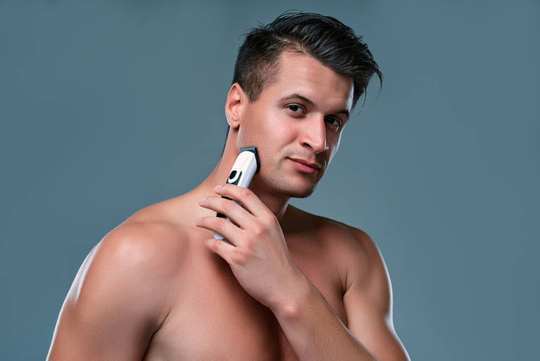 Handsome young man isolated. Portrait of shirtless muscular man is standing on gray background with trimmer in hand while shaving. Man care concept. - Photo, Image