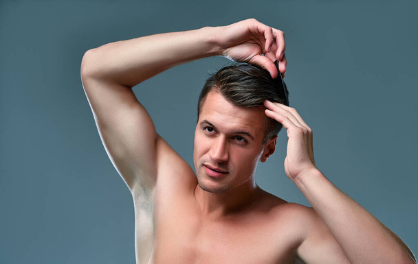 Un joven apuesto aislado. Retrato de un hombre musculoso sin camisa está de pie sobre un fondo gris y peinando su cabello. - Foto, Imagen