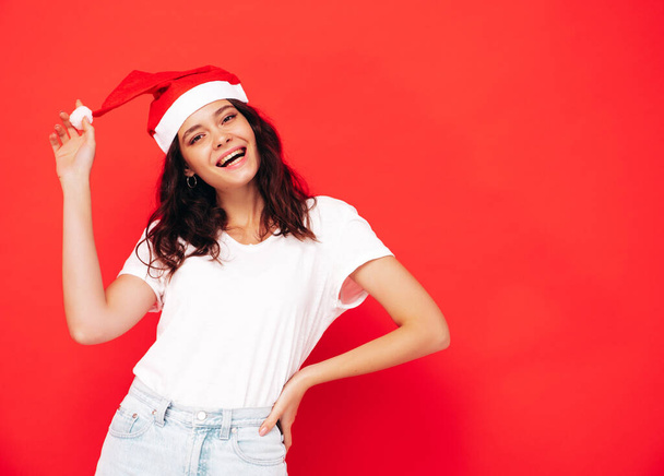 Beautiful woman celebrating New Year.Happy gorgeous female in casual clothes having fun at New Year's Eve party.Holiday celebration.Charming model in Santa Claus hat laughing near red wall - Foto, Imagem