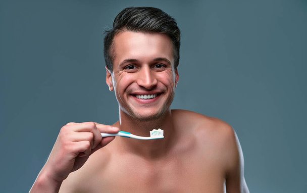 Beau jeune homme isolé. Portrait de l'homme musclé torse nu est debout sur fond gris avec brosse à dents et dentifrice à la main et souriant. Hommes concept de soins
. - Photo, image