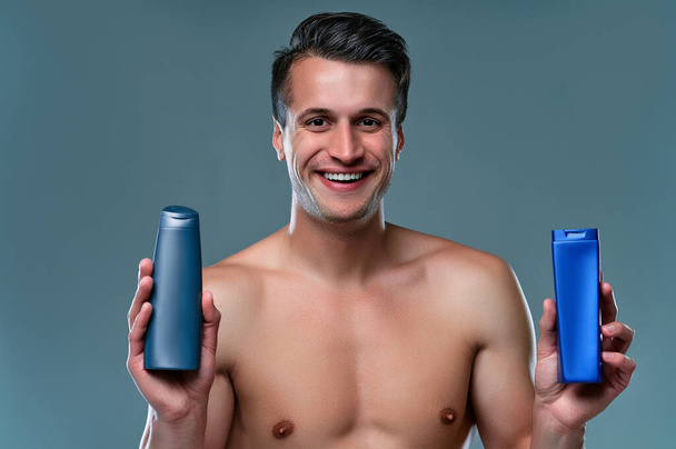 Handsome young man isolated. Portrait of shirtless muscular man is standing on grey background with shampoo in hands. Men care concept. - Photo, Image