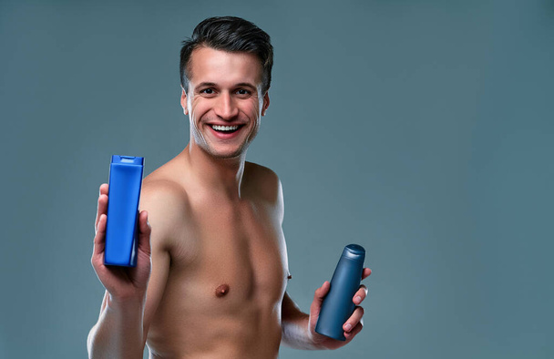 Handsome young man isolated. Portrait of shirtless muscular man is standing on grey background with shampoo in hands. Men care concept. - Photo, Image
