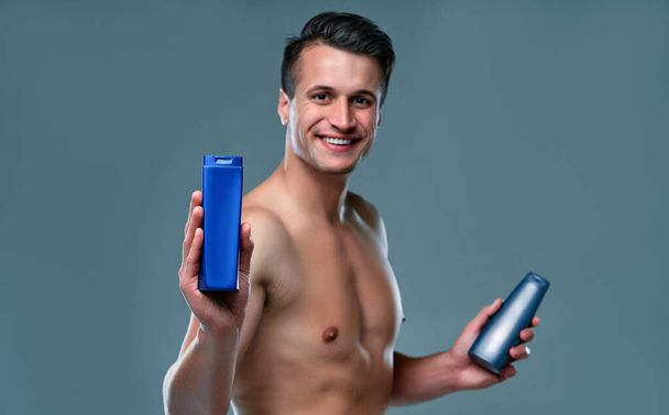 Handsome young man isolated. Portrait of shirtless muscular man is standing on grey background with shampoo in hands. Men care concept. - Zdjęcie, obraz