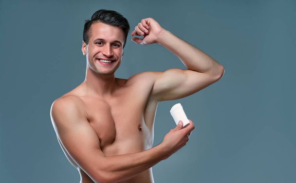 Handsome young man isolated. Portrait of shirtless muscular man is standing on grey background and using antiperspirant. Men care concept. - Fotoğraf, Görsel