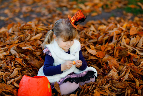 Маленькая девочка, одетая как ведьмин трюк или угощение на Хэллоуин. Счастливый ребенок на улице, в оранжевой веселой шляпе и мешке для сладкого. Сезон семейных фестивалей в октябре. Активность на открытом воздухе - Фото, изображение